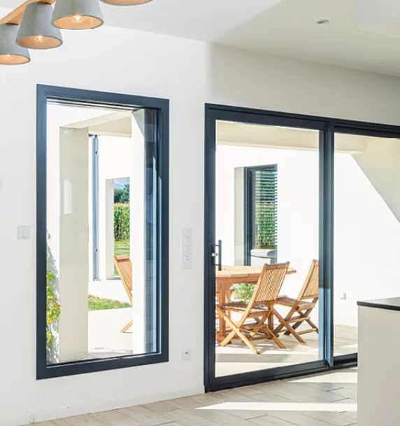 Une porte vitrée noire s'ouvre sur une terrasse ensoleillée avec une table et des chaises en bois, dans une maison moderne.