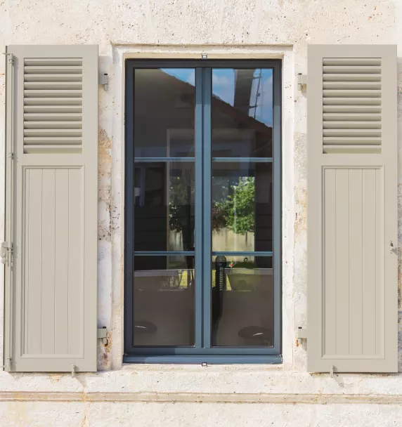 Une fenêtre à volets gris ouverts expose une vue extérieure sur un jardin, entourée d'un mur en pierre beige.