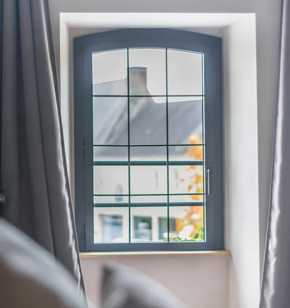 Une fenêtre à croisillons, encadrant une vue sur des toits et des arbres colorés d'automne, est bordée de rideaux gris ouverts.