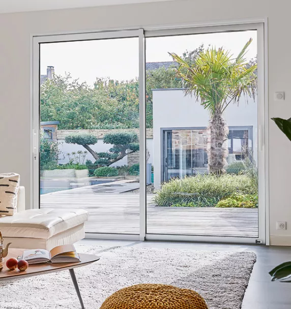 Une maison moderne avec une grande baie vitrée s'ouvre sur un jardin verdoyant avec une terrasse en bois, une palmeraie et des buissons.