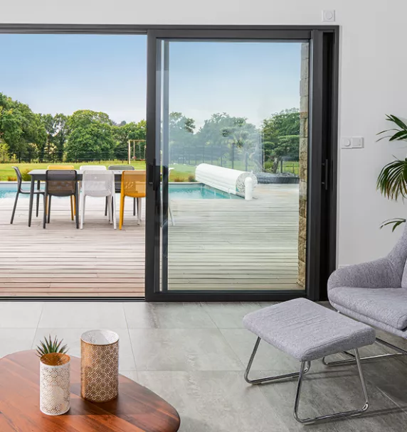 Salon moderne avec vue sur une terrasse en bois, donnant sur une piscine et un jardin verdoyant. Fauteuil et table basse agrémentent l'espace intérieur.