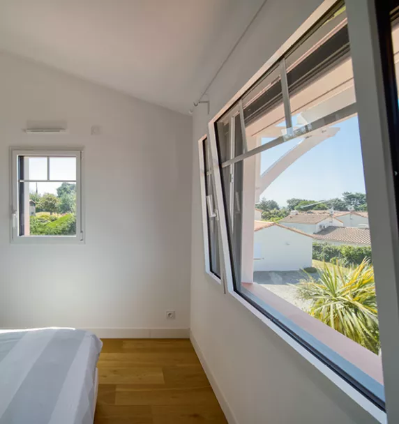 Fenêtre ouverte sur mur blanc, laissant entrer la lumière et offrant une vue sur des toits de maisons et des arbres, dans une chambre aux murs blancs.