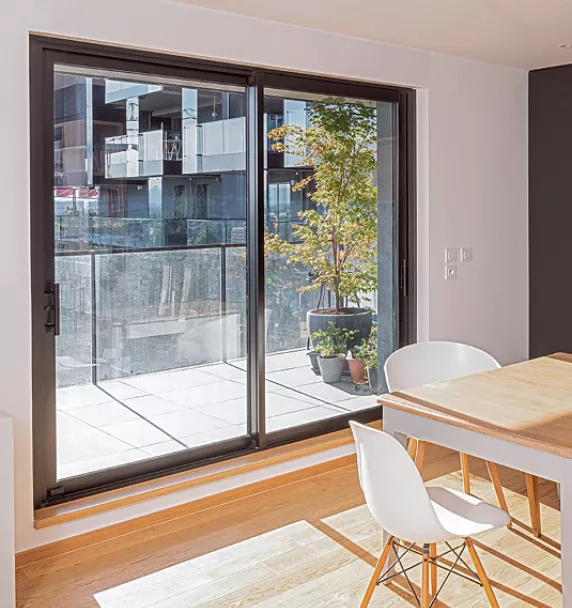 Grande baie vitrée donnant sur un balcon avec des plantes en pot, située dans une pièce à manger lumineuse avec une table et des chaises blanches.