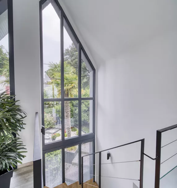 Grande fenêtre vitrée laisse entrer la lumière naturelle, illuminant un escalier moderne avec des plantes et une vue sur un jardin verdoyant.
