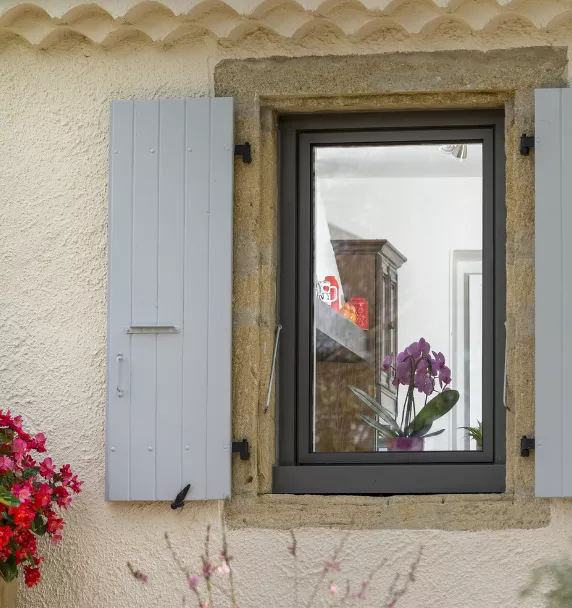 Une fenêtre ouverte, encadrée de volets bleus, dévoile à l'intérieur une orchidée violette sur une étagère.