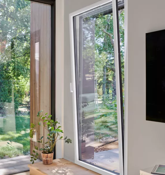 Fenêtre basculée laissant entrer la lumière, avec un pot de plante à côté, donnant sur un jardin verdoyant.