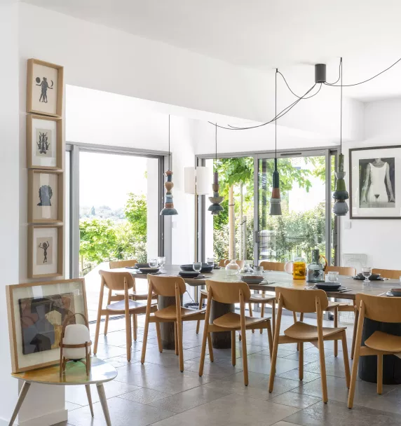 Table en bois entourée de chaises, décorée avec des éléments de vaisselle, dans une salle à manger lumineuse avec de larges baies vitrées donnant sur un jardin verdoyant.
