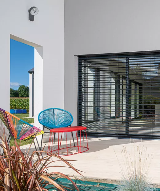 Mobilier coloré, placé sur une terrasse moderne, accompagné de stores en lamelles, avec une vue dégagée sur le jardin et le ciel bleu.
