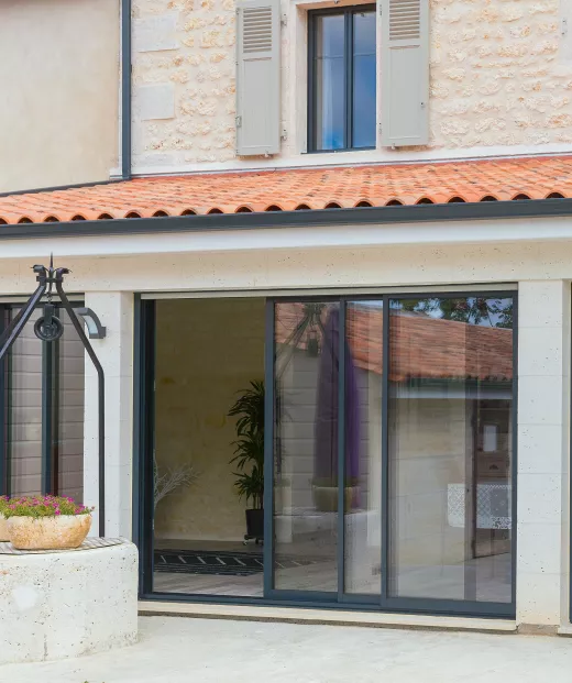 Porte-fenêtre vitrée ouvre sur terrasse ensoleillée, entourée de murs en pierre claire et d'une plante en pot à l'intérieur.