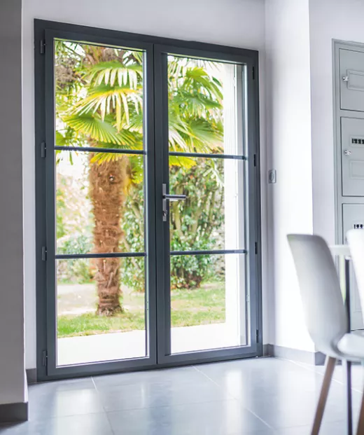 Porte vitrée double, donnant sur un jardin arboré avec palmiers, située dans une pièce lumineuse et moderne avec des murs blancs et un sol en carrelage gris.