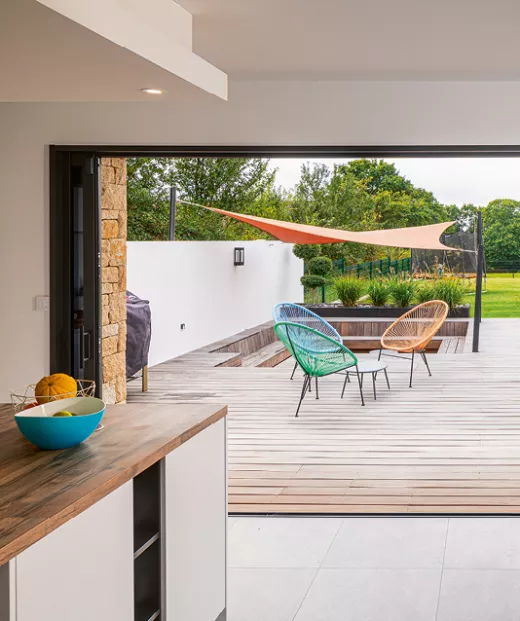 Chaises colorées disposées sur une terrasse extérieure en bois, abritées par une voile d'ombrage, donnent sur un jardin verdoyant.
