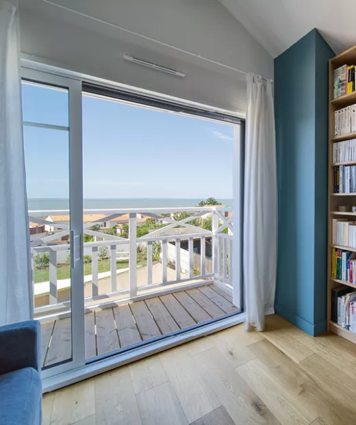 Une grande baie à galandage vitrée ouverte donnant sur un balcon en bois, avec vue sur la mer et des toits de maisons proches, intégrée à un salon lumineux avec des étagères remplies de livres.