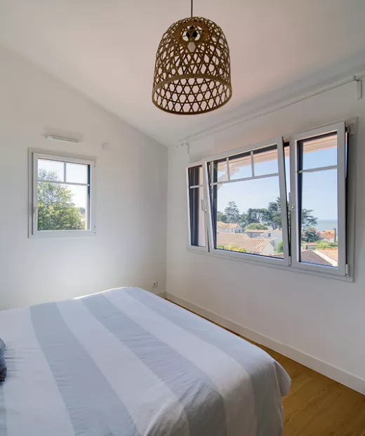 Lit d’une chambre minimaliste, éclairé par des fenêtres ouvertes donnant sur une vue arborée et la mer, sous une suspension en rotin.