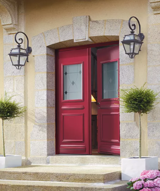 Une porte d'entrée rouge ouverte, encadrée par des murs en pierre, avec deux lanternes noires et des arbustes en pots de chaque côté.