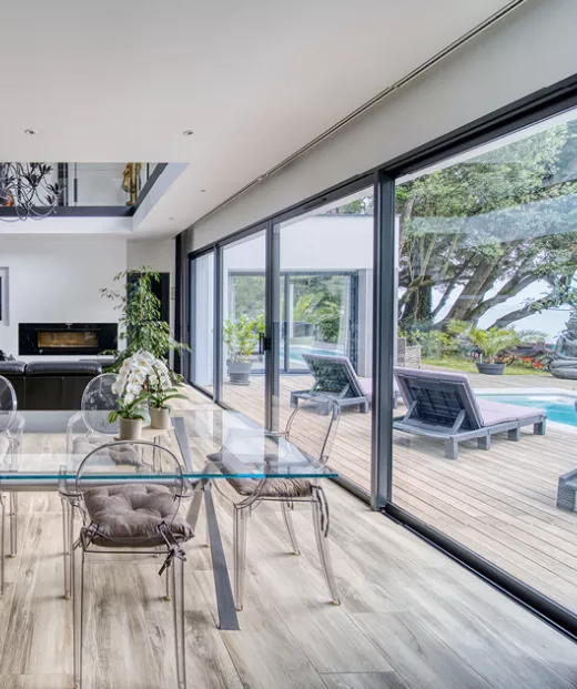 Table en verre, entourée de chaises transparentes, placée dans un salon moderne avec vue sur une terrasse en bois et une piscine extérieure.