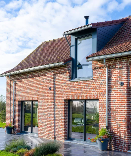 Maison en briques rouges, avec grandes fenêtres et un toit en tuiles, située dans un jardin verdoyant sous un ciel légèrement nuageux.