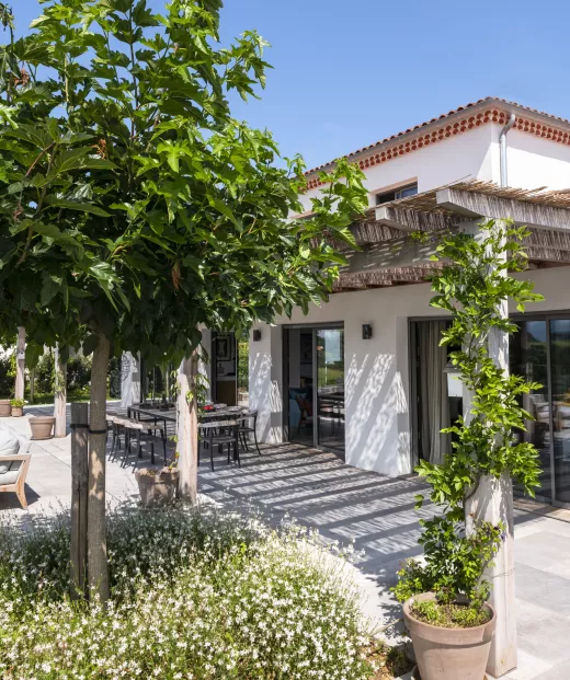 Maison avec terrasse ombragée, entourée de plantes vertes et fleurs blanches, située sous un ciel bleu clair. Tables et chaises en métal sont disposées pour des repas en plein air.