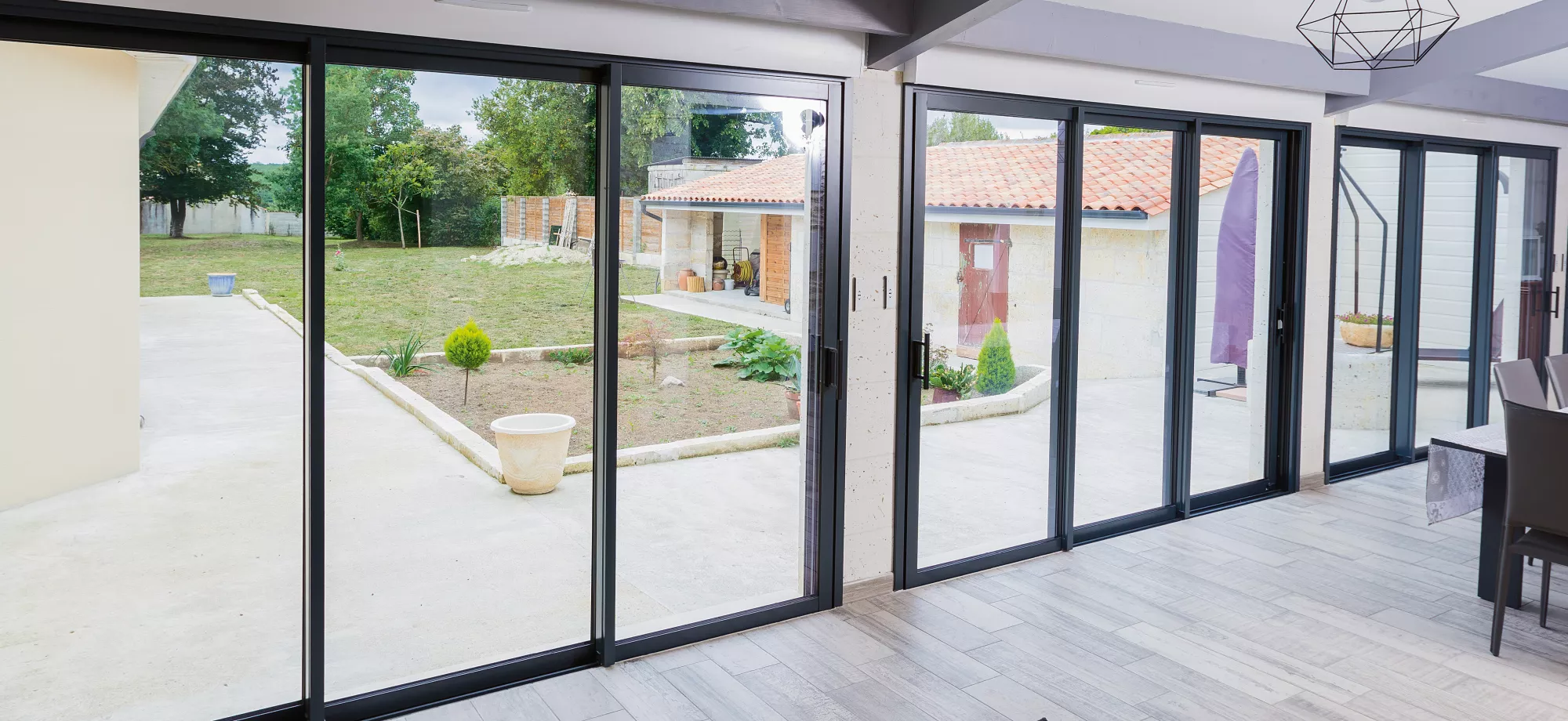 De grandes baies vitrées coulissantes offrent une vue sur un jardin aménagé, intégrant des bâtiments avec tuiles rouges en arrière-plan.