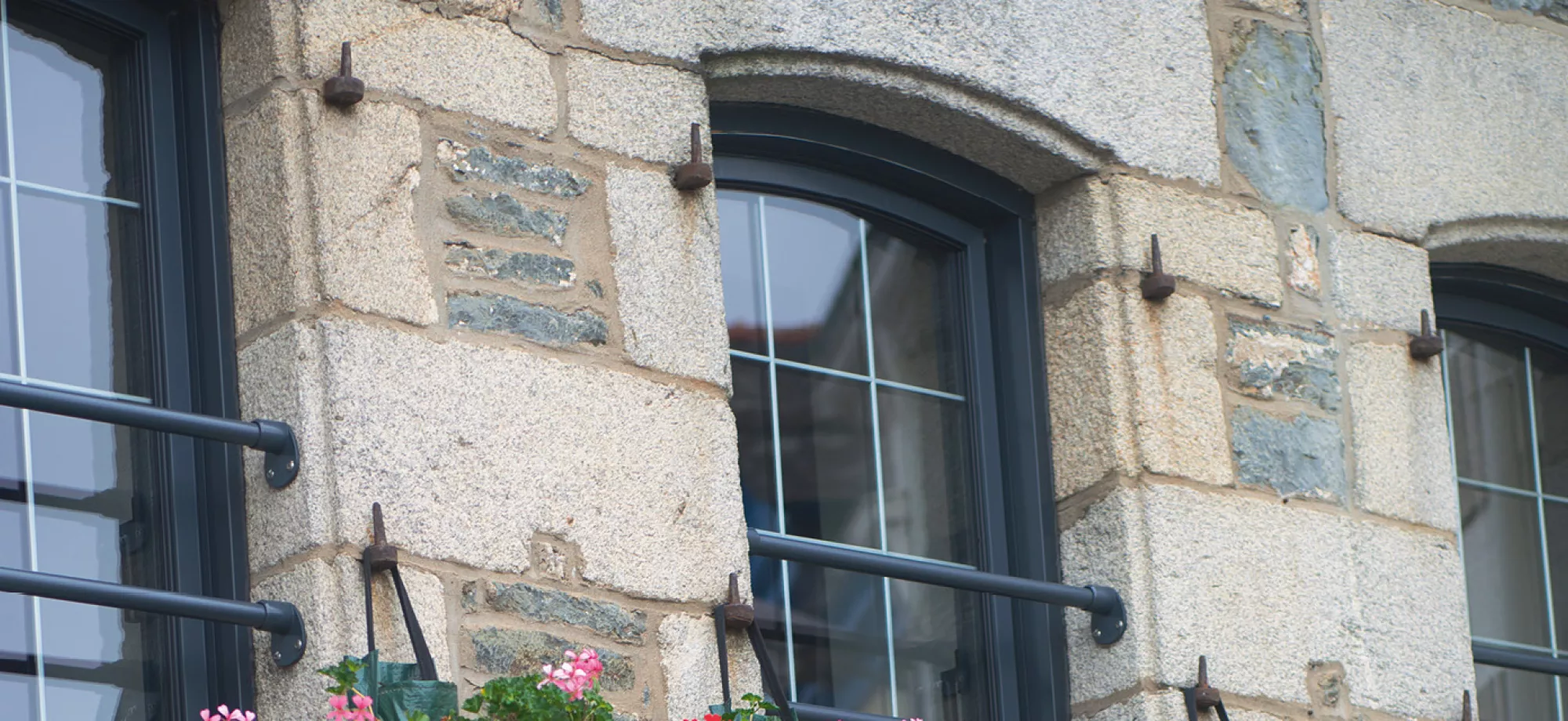 Une fenêtre à encadrement noir avec barreaux, ornée de fleurs colorées, est intégrée dans un mur en pierre brute dans un environnement urbain.
