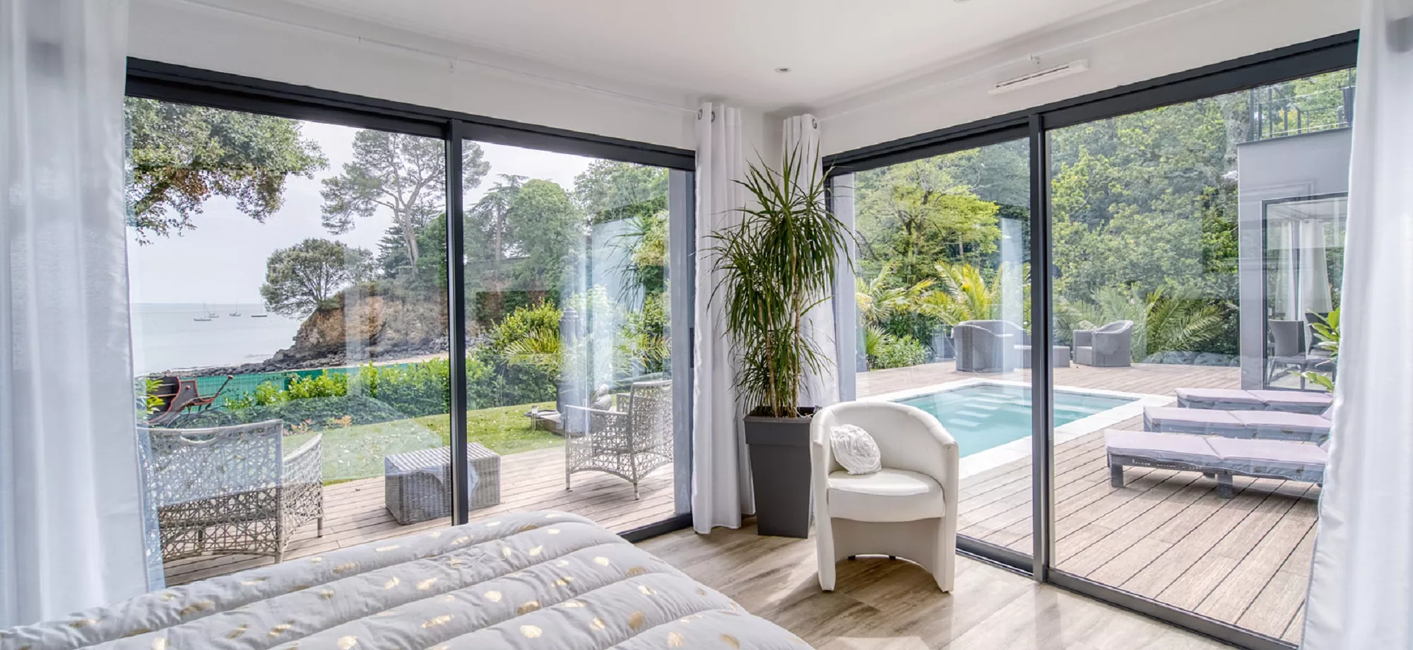 Une chambre lumineuse avec grandes baies vitrées donnant sur une terrasse en bois, une piscine, et un jardin avec vue sur la mer.