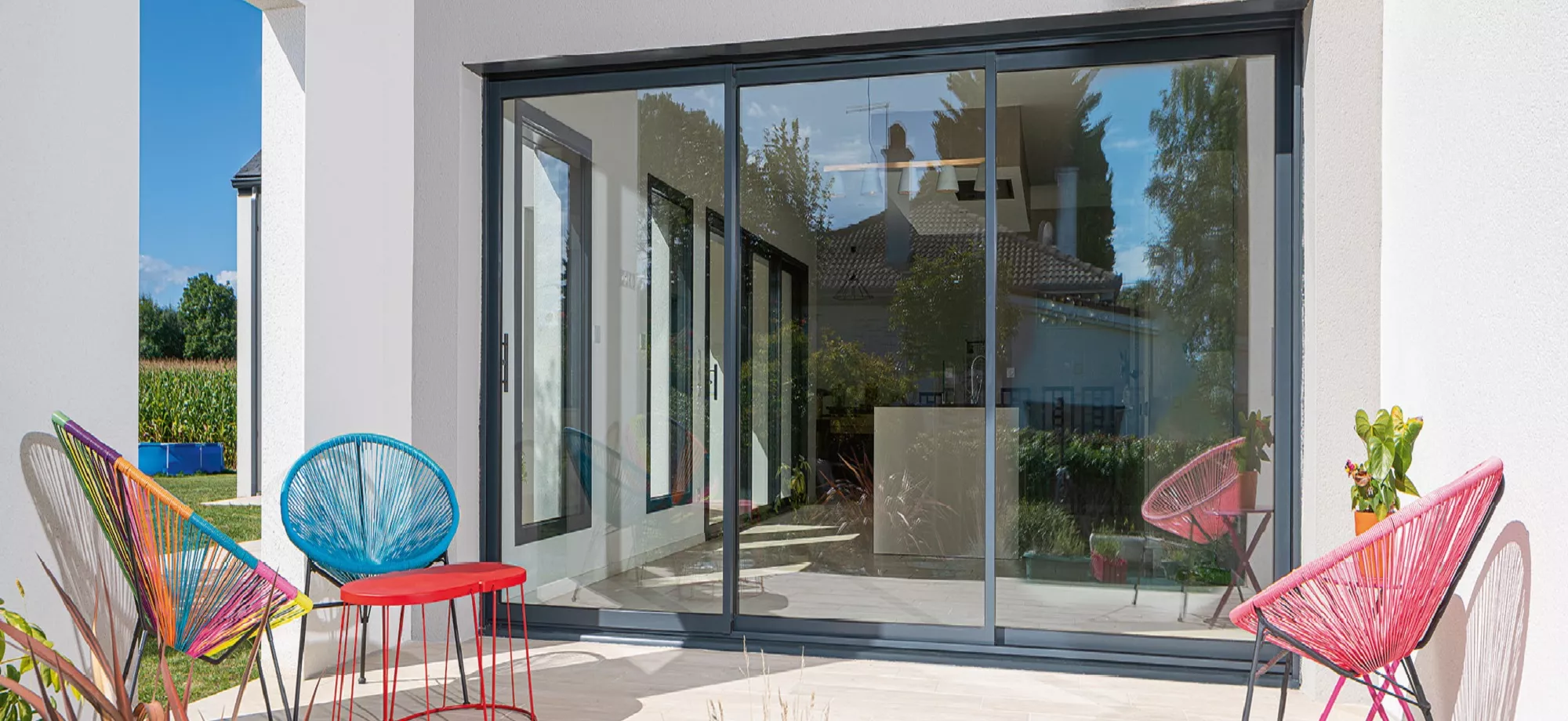 Chaises colorées entourant une table rouge, placées sur une terrasse ensoleillée devant des portes vitrées donnant sur un jardin.
