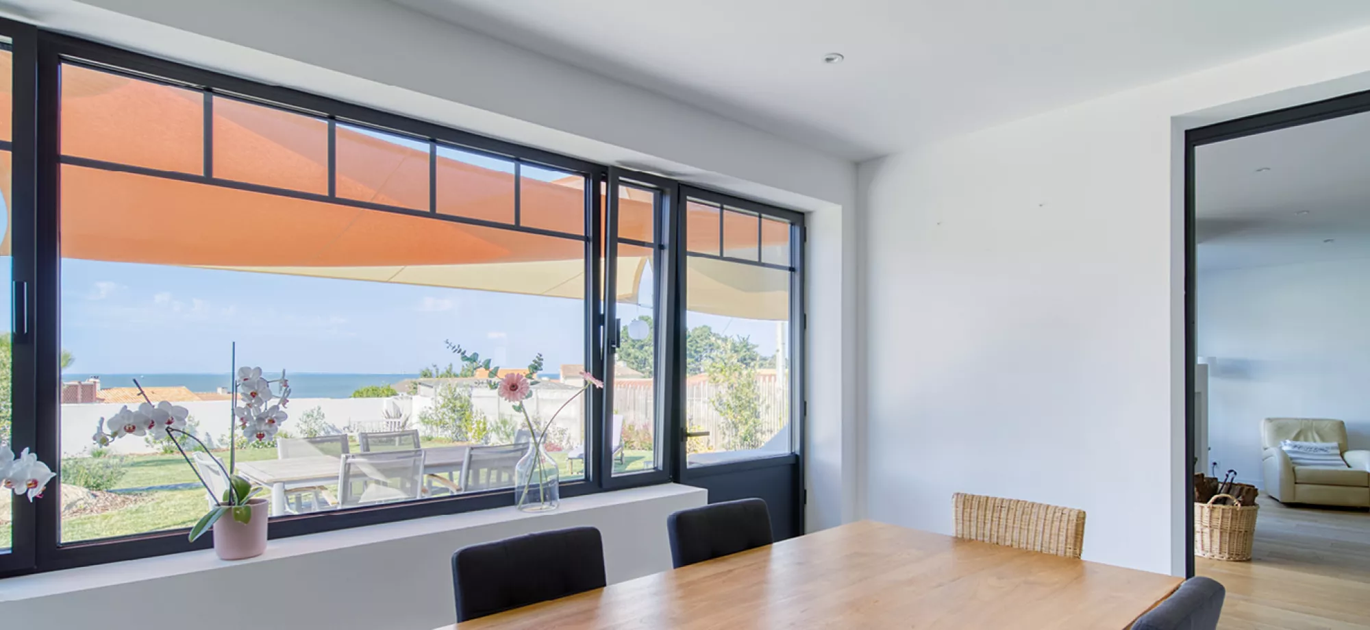 Table en bois entourée de chaises, fleur en pot près de la fenêtre ouverte sur terrasse et vue sur la mer.