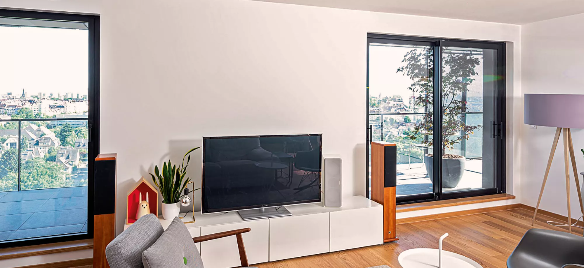 Télévision éteinte, posée sur un meuble bas blanc, jouxtant des enceintes en bois, dans un salon moderne avec vue dégagée sur la ville à travers grandes baies vitrées.