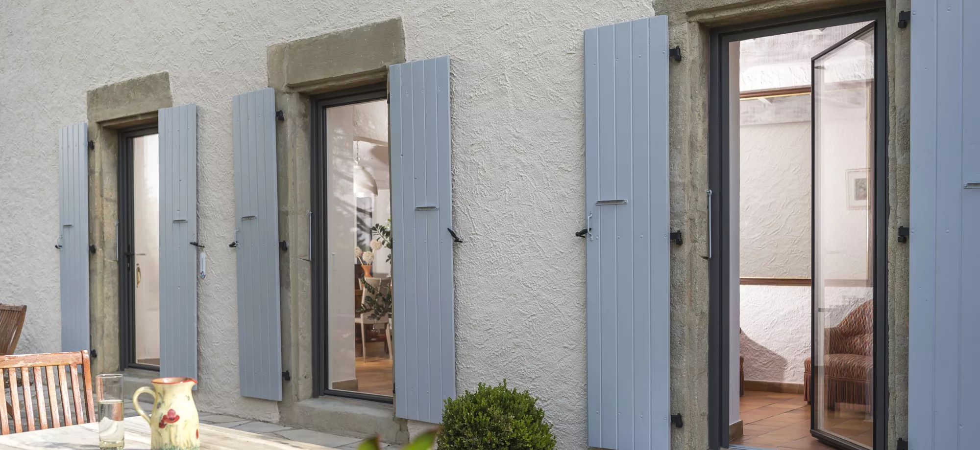Volets bleus encadrent des portes-fenêtres ouvertes, laissant entrevoir un intérieur lumineux avec des plantes, devant une terrasse avec une table en bois et une carafe.