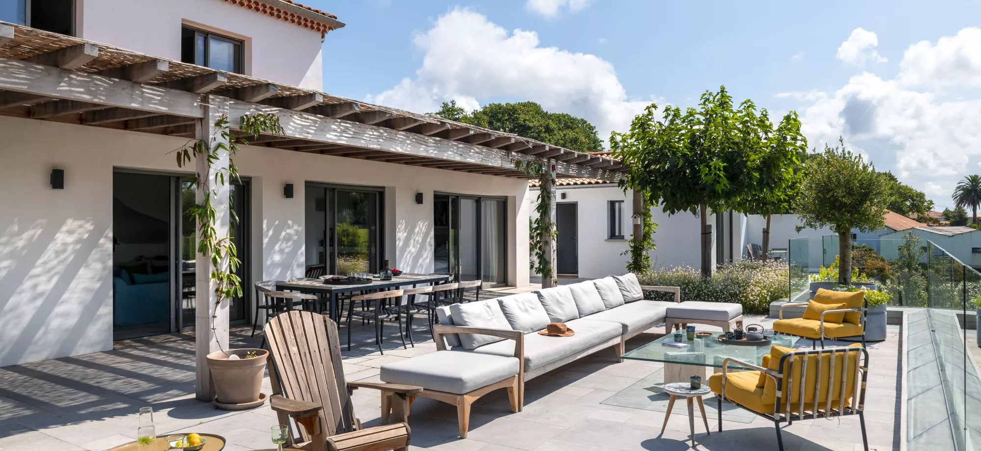 Salon de jardin avec meubles en bois sous une pergola, entouré de plantes et d'arbres sur une terrasse ensoleillée.