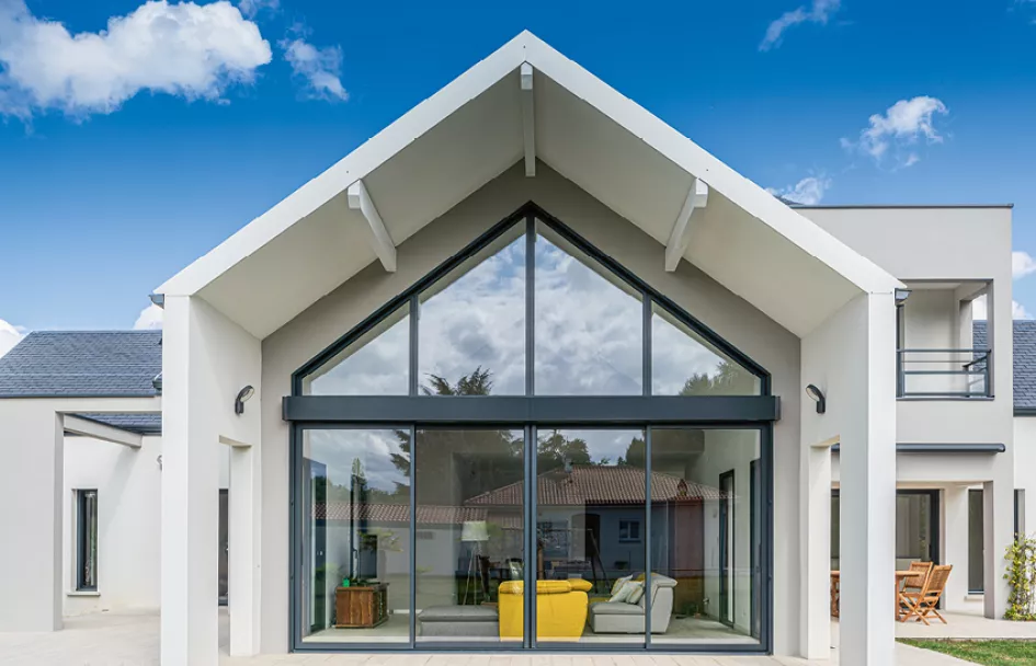 Maison moderne avec grandes baies vitrées, située dans un environnement résidentiel, reflétant les arbres et le ciel bleu par temps ensoleillé. Deux fauteuils sont visibles à l'intérieur.