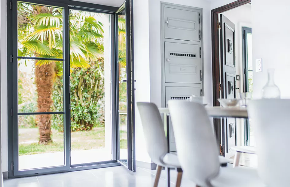 Porte-fenêtre ouverte donnant sur un jardin verdoyant, vue depuis une salle à manger moderne avec table et chaises blanches.