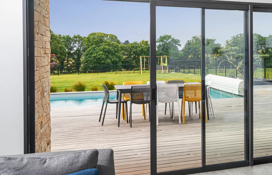 Table entourée de chaises colorées, posée sur une terrasse en bois, près d'une piscine, avec un jardin verdoyant et une balançoire en arrière-plan.