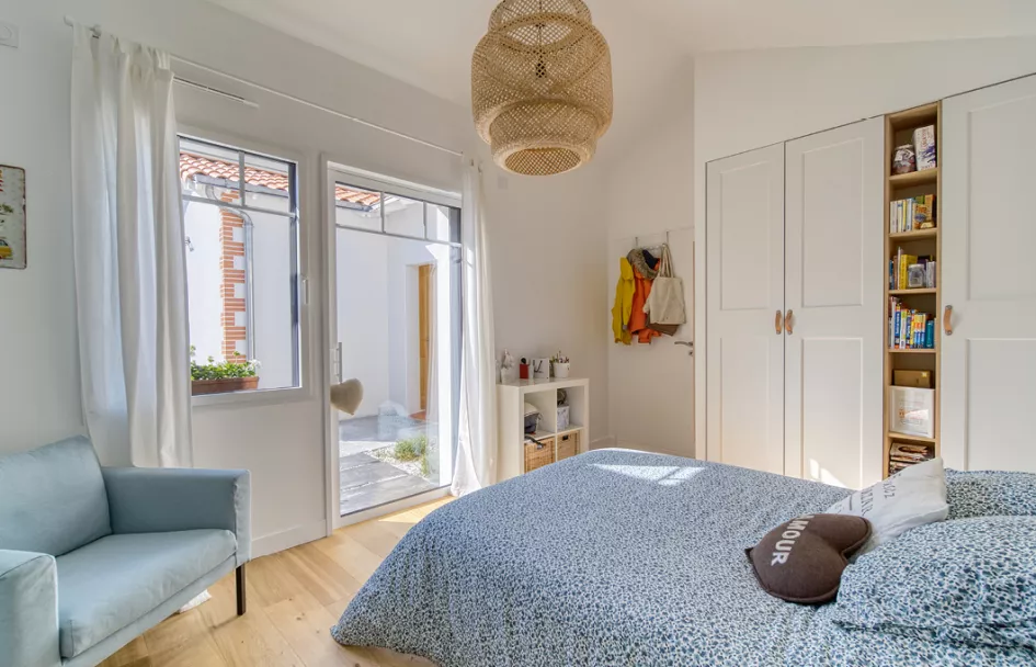 Une chambre lumineuse avec un lit, une armoire blanche, une étagère et une porte-fenêtre donnant sur une cour pavée.