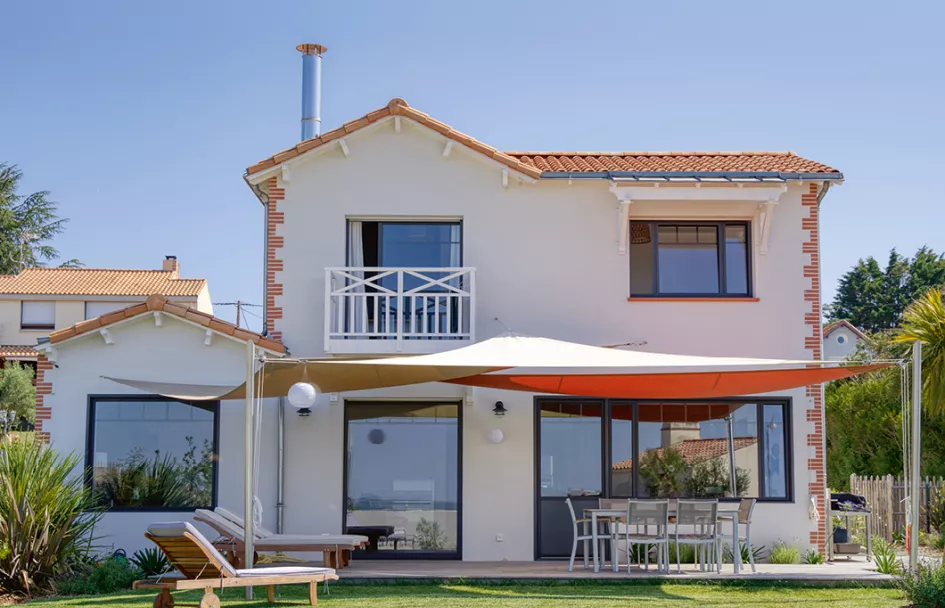 Maison blanche à deux étages avec toit en tuiles rouges, équipées de chaises longues et table sur une terrasse dans un jardin, avec un auvent beige.