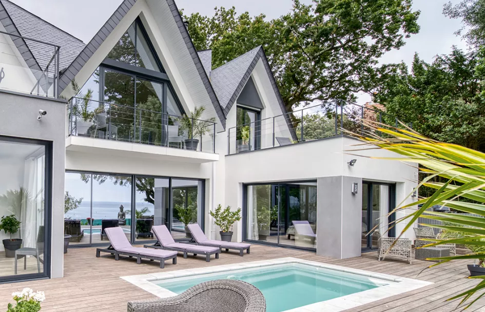 Maison moderne avec grandes fenêtres et terrasse, des transats autour d'une petite piscine, située dans un environnement verdoyant proche de la mer.