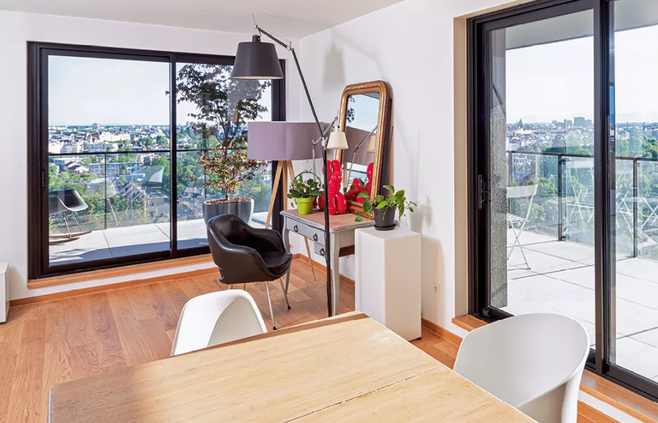 Un salon lumineux avec des fauteuils modernes et une table, donnant sur un balcon avec vue panoramique sur la ville, grâce à de grandes baies vitrées.