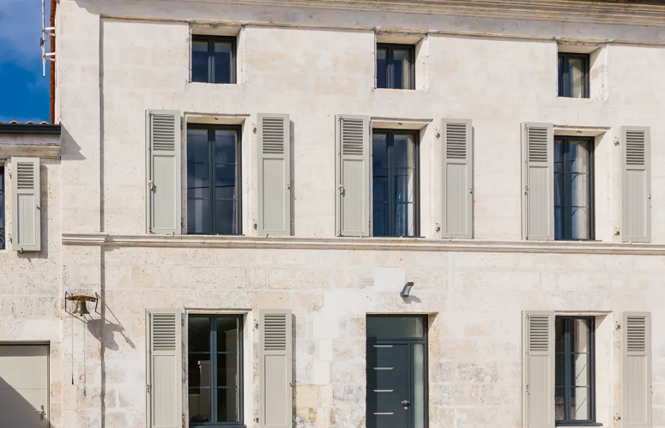 Une façade de maison en pierre avec des volets gris fermés, baignée par une lumière ensoleillée, donnant une apparence calme et ordonnée.