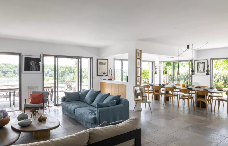 Salon moderne avec canapés bleus, donnant sur une salle à manger lumineuse et de grandes baies vitrées offrant une vue sur la nature extérieure.