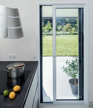 Porte-fenêtre vitrée s'ouvrant sur un jardin verdoyant, avec une plante en pot à droite. Cuisine moderne avec casserole sur une plaque et des fruits sur le comptoir.