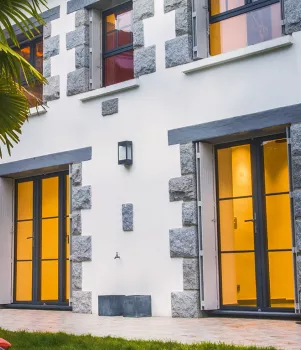 Maison moderne, éclairée, avec façades en pierres grises et fenêtres grandes, se trouve dans un jardin verdoyant à côté d'un palmier et des fauteuils en métal.