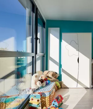 Des jouets en peluche reposent près d'une fenêtre lumineuse, dans une chambre d'enfant avec une armoire blanche et un mur turquoise.