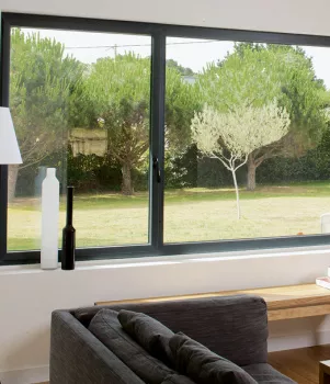 Un canapé gris est placé dans un salon lumineux avec une grande fenêtre qui donne sur un jardin verdoyant.