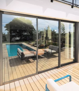Une large baie vitrée reflète une terrasse en bois avec piscine, entourée de chaises et arbustes en arrière-plan.