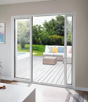 Une grande porte-fenêtre coulissante s'ouvre sur une terrasse en bois, où un canapé et une table basse sont placés, donnant sur un jardin verdoyant.