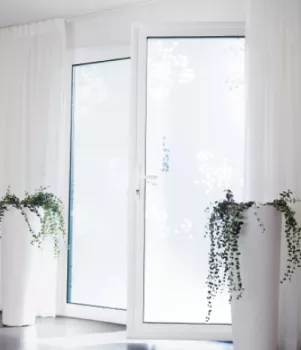 Porte-fenêtre vitrée, décorée de motifs flocons, encadrée de rideaux blancs, éclairée par la lumière naturelle et ornée de deux plantes en pots blancs des deux côtés.