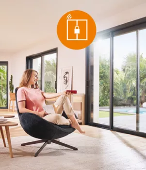 Une femme assise dans un fauteuil utilise une télécommande pour ouvrir une baie vitrée, donnant sur un jardin ensoleillé avec piscine.