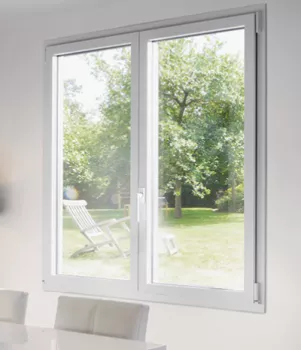 Une grande fenêtre blanche laisse voir un jardin verdoyant avec des arbres et des chaises longues blanches, baigné de lumière naturelle.