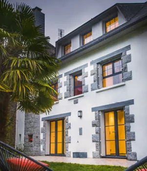 Maison en pierre blanche avec fenêtres lumineuses, entourée d'un jardin verdoyant, montrant un palmier.