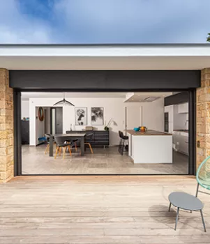 Une terrasse en bois jouxtant une cuisine et une salle à manger ouvertes, avec mobilier moderne, visibles à travers une grande baie vitrée.
