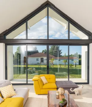 Un salon lumineux avec de grands canapés jaunes, donnant sur un jardin à travers d'immenses baies vitrées, avec un chat marchant devant une table basse en bois.